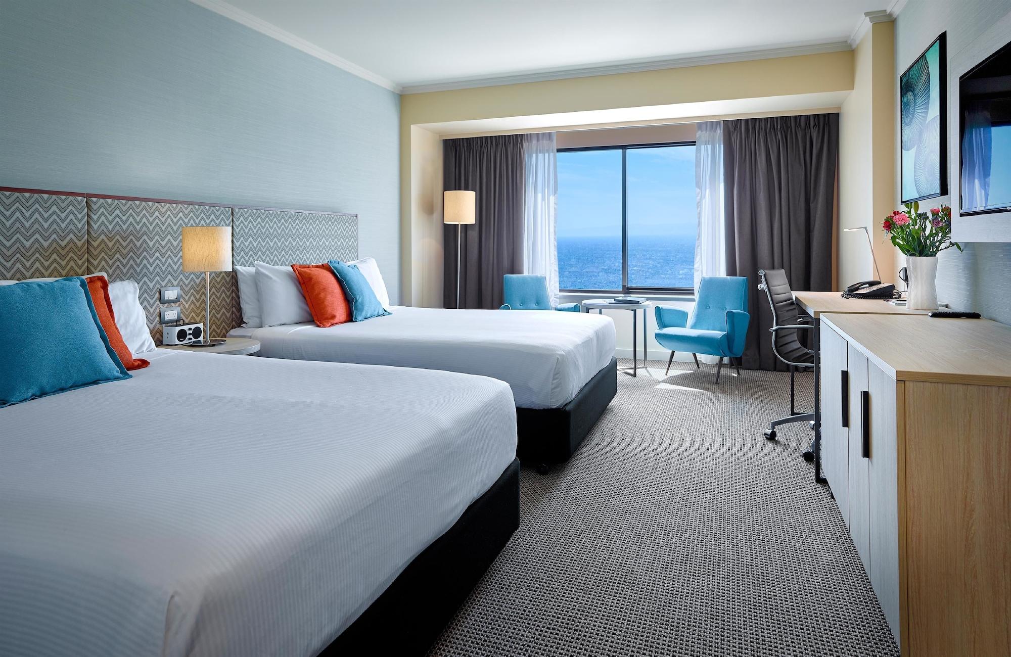 Отель Stamford Grand Adelaide Гленелг Экстерьер фото The photo shows a well-decorated hotel room featuring two queen-sized beds with white linens and colorful accent pillows. There is a large window with curtains that offers a view of the ocean. A small seating area with a blue chair and a matching des