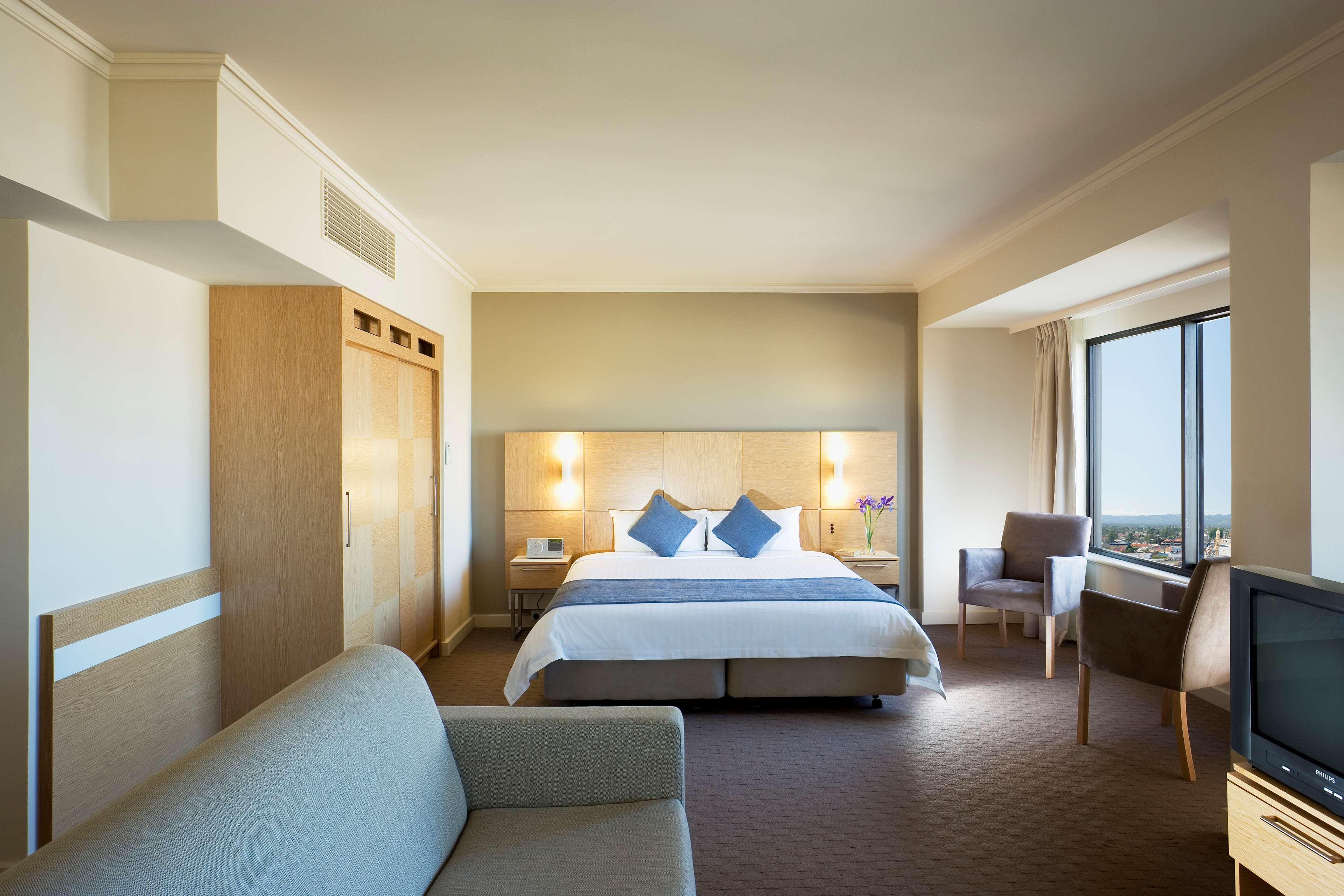 Отель Stamford Grand Adelaide Гленелг Номер фото The photo shows a modern hotel room featuring a large bed with two blue decorative pillows. There are bedside lamps on both sides of the headboard. To the left, there's a wooden wardrobe and a small table with a vase and a few amenities. On the right