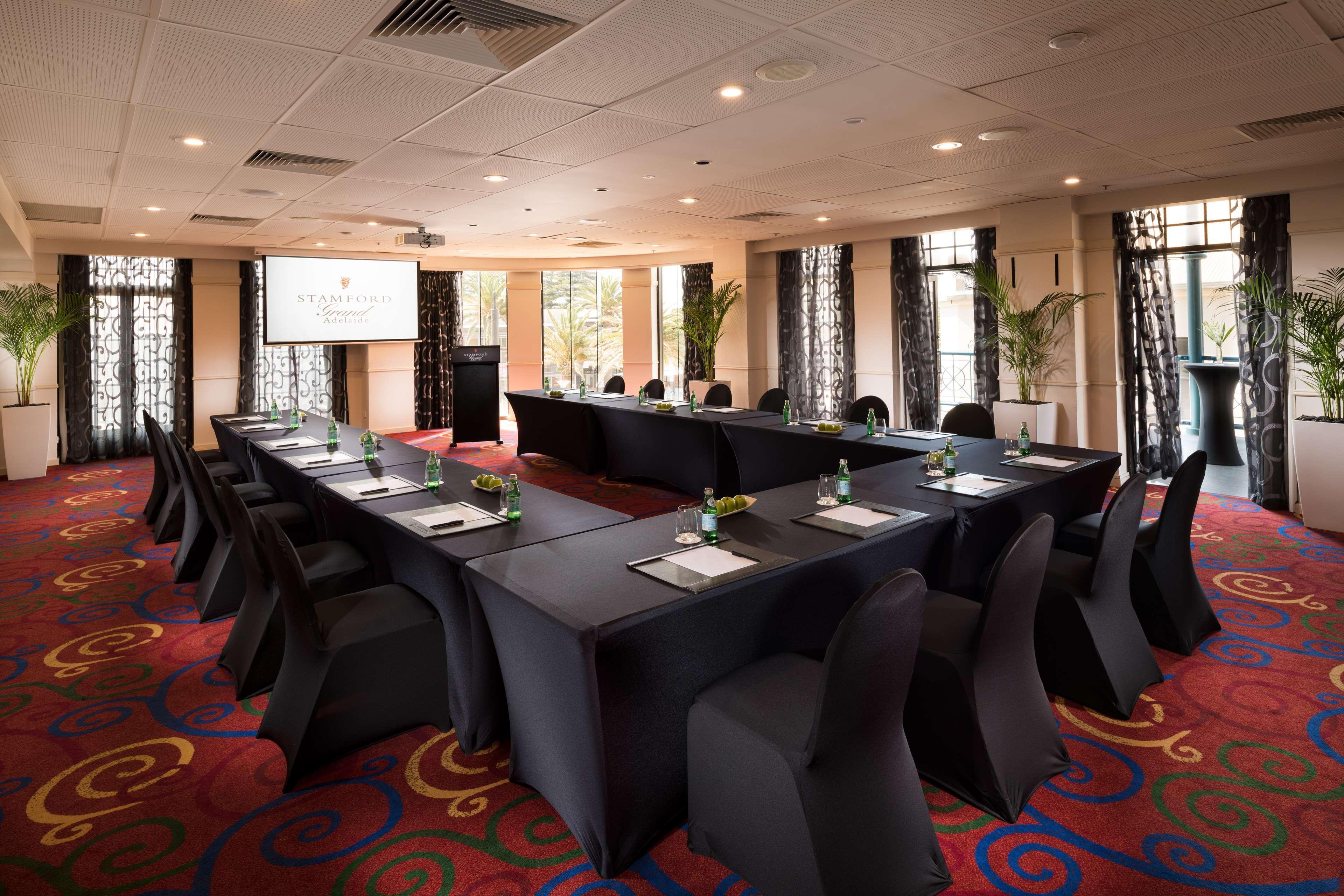 Отель Stamford Grand Adelaide Гленелг Экстерьер фото The photo shows a conference room set up for a meeting or presentation. There are several tables arranged in a U-shape, covered with black tablecloths. Each table has a notepad and a small bottle of water on it. The chairs around the tables are also 