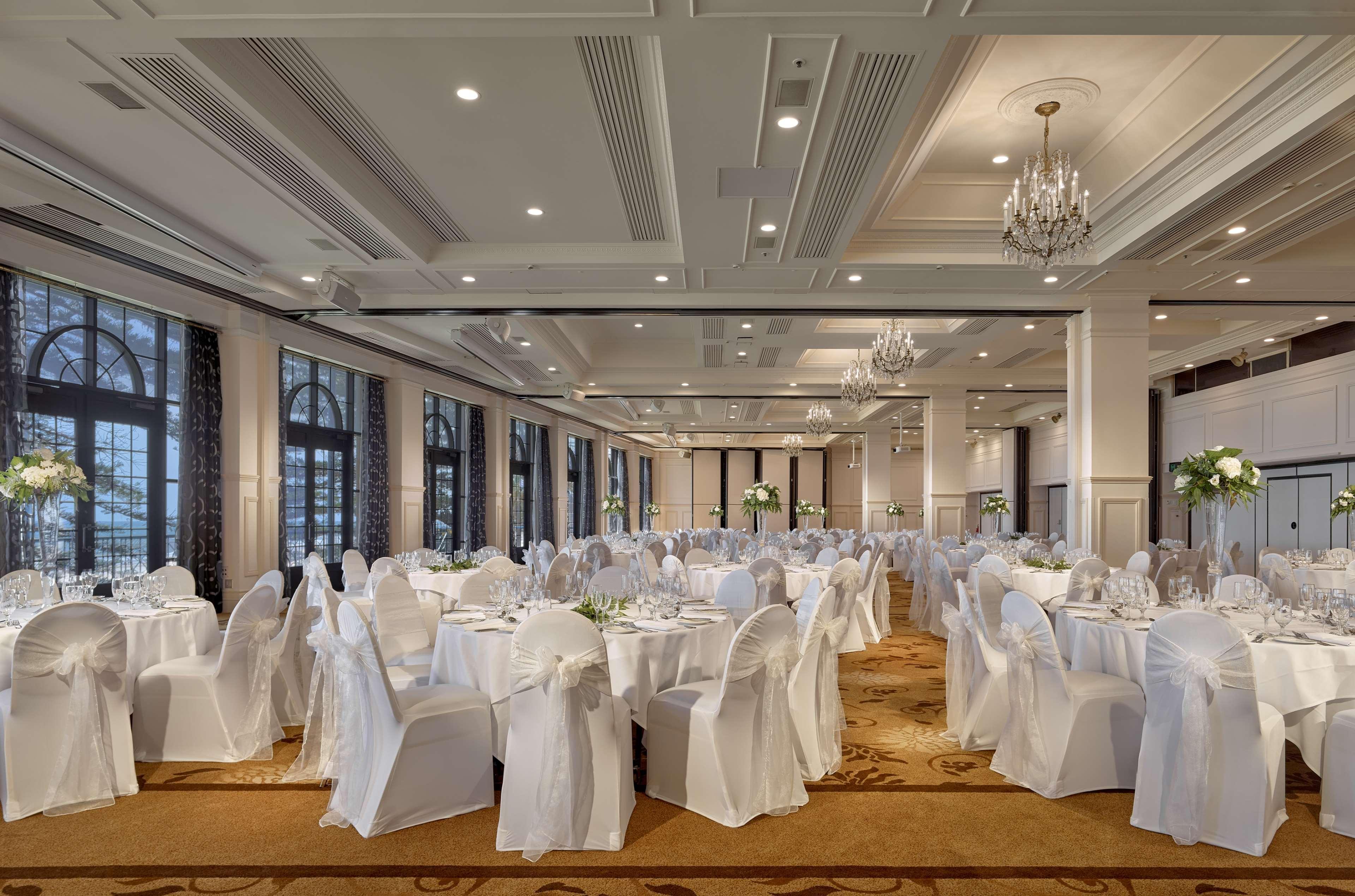 Отель Stamford Grand Adelaide Гленелг Экстерьер фото The photo depicts an elegantly arranged banquet hall or dining area. The space features multiple round tables, each set with white tablecloths and decorated with floral centerpieces. The chairs are covered in white fabric, contributing to a formal at