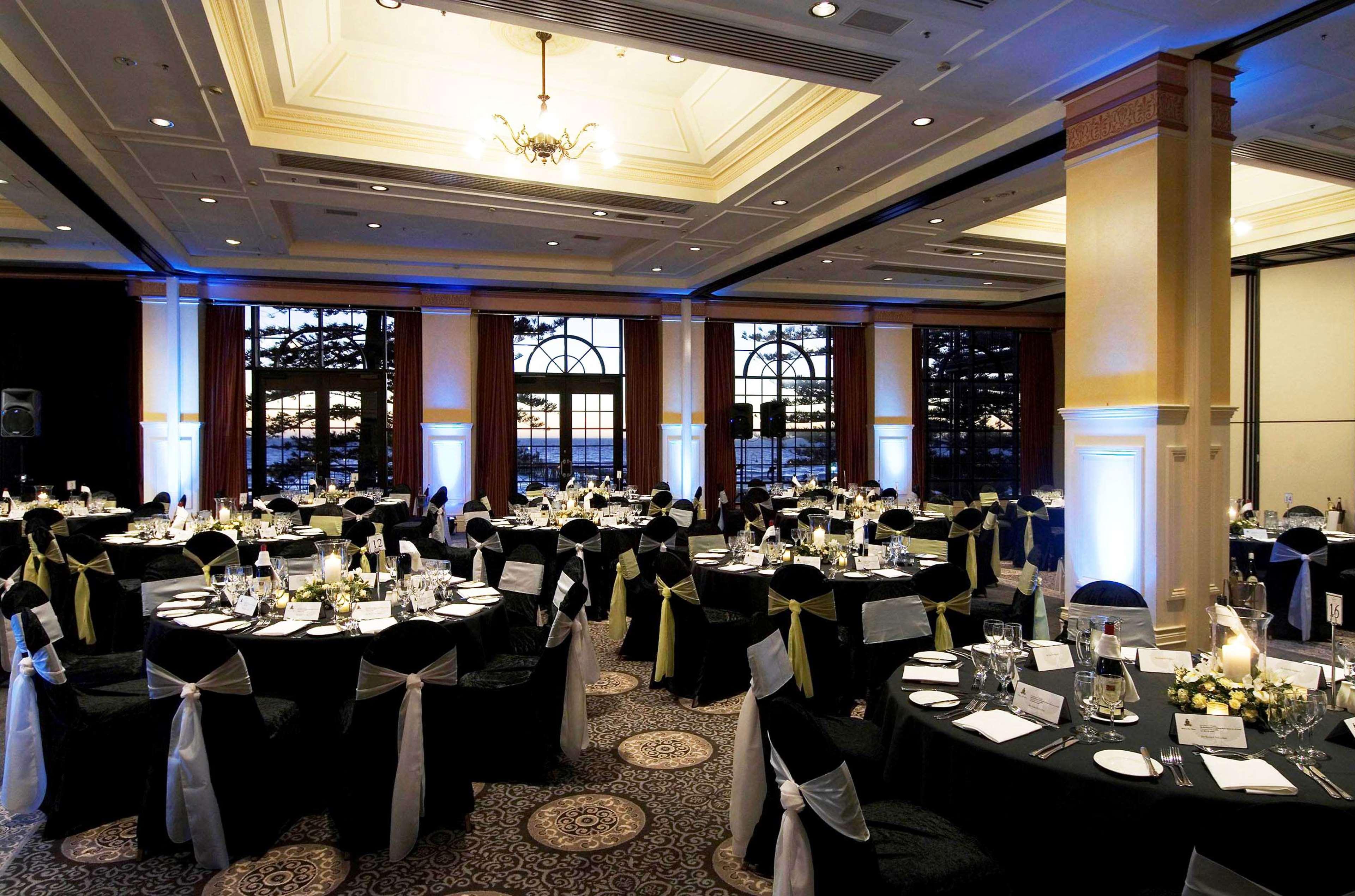 Отель Stamford Grand Adelaide Гленелг Ресторан фото The photo depicts an elegantly arranged banquet hall set for an event. The room features round tables covered with black tablecloths, each adorned with white tableware and floral centerpieces. Chairs are draped in white fabric with decorative sashes 