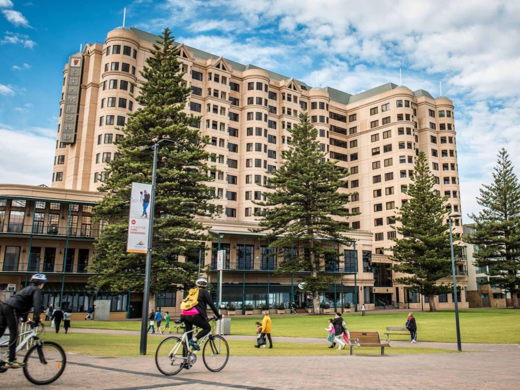 Отель Stamford Grand Adelaide Гленелг Экстерьер фото The photo depicts a modern building with a large facade, likely a hotel or commercial establishment, situated near a spacious green area. Surrounding the building are several tall trees, providing shade and enhancing the aesthetic appeal. In the fore