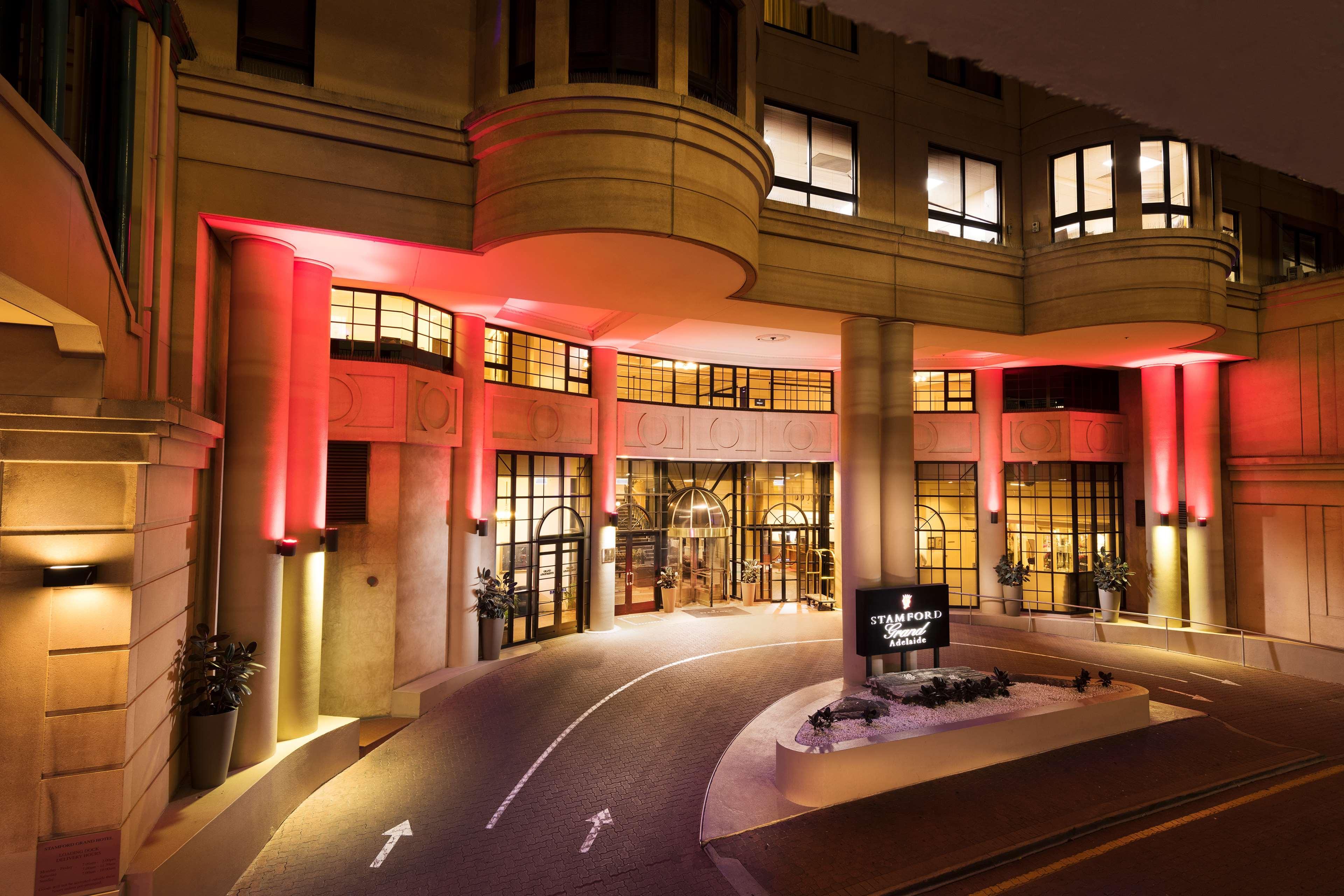 Отель Stamford Grand Adelaide Гленелг Экстерьер фото The photo shows the entrance of a hotel or luxury building at night. The façade features a modern architectural style with large windows and an elegant entrance. The area is illuminated with red lighting, creating an inviting atmosphere. There appear