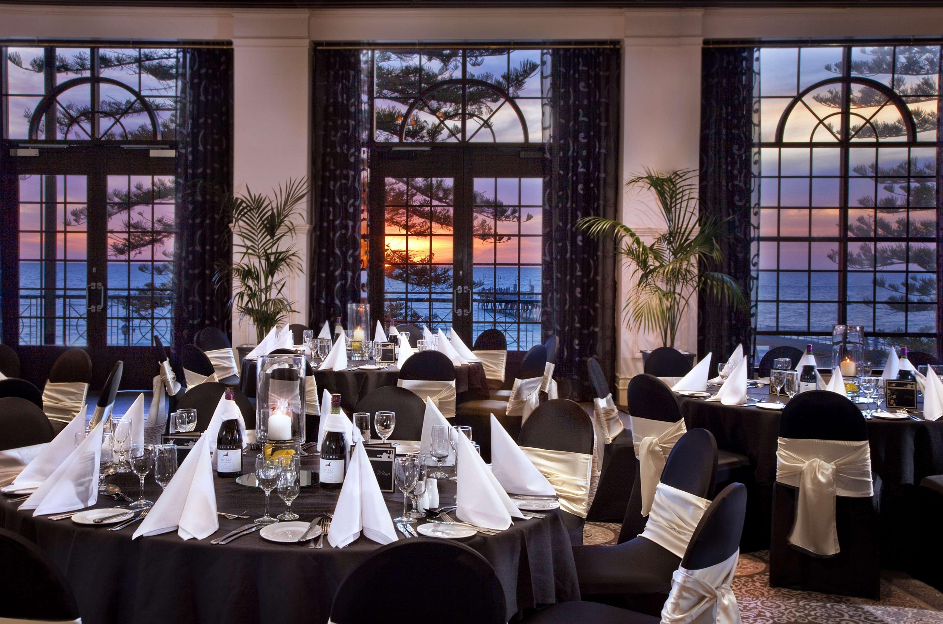 Отель Stamford Grand Adelaide Гленелг Ресторан фото The photo shows an elegantly decorated dining room with a beautiful view. The tables are set with black tablecloths and white napkins folded in a triangular shape. There are glassware and silverware neatly arranged at each setting. The room is adorne