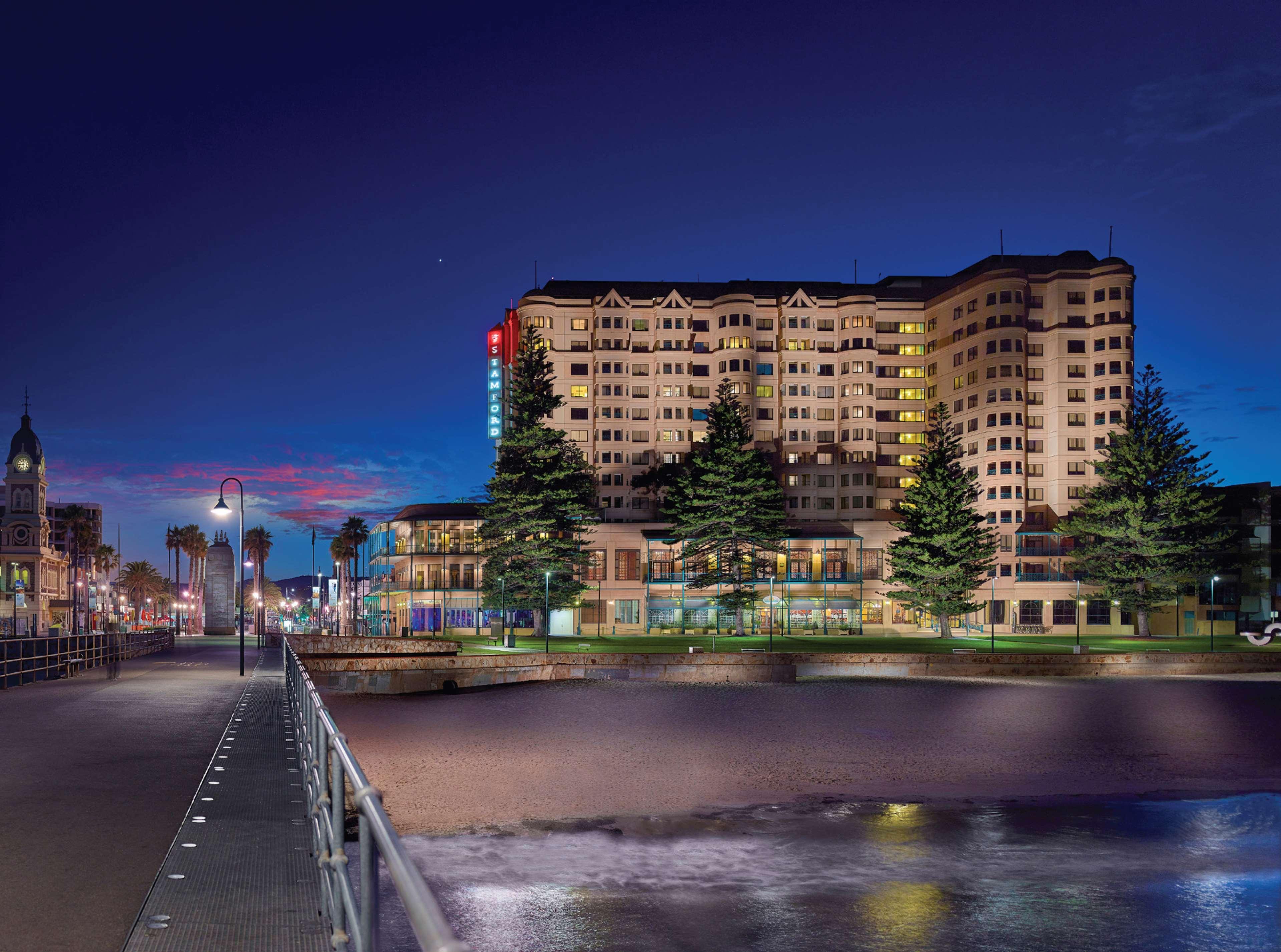 Отель Stamford Grand Adelaide Гленелг Экстерьер фото The photo shows a waterfront scene at dusk, featuring a large building lit up against a darkening sky. The architecture appears modern and prominent, with several windows illuminated. In the foreground, there is a walkway or pier with fencing along t