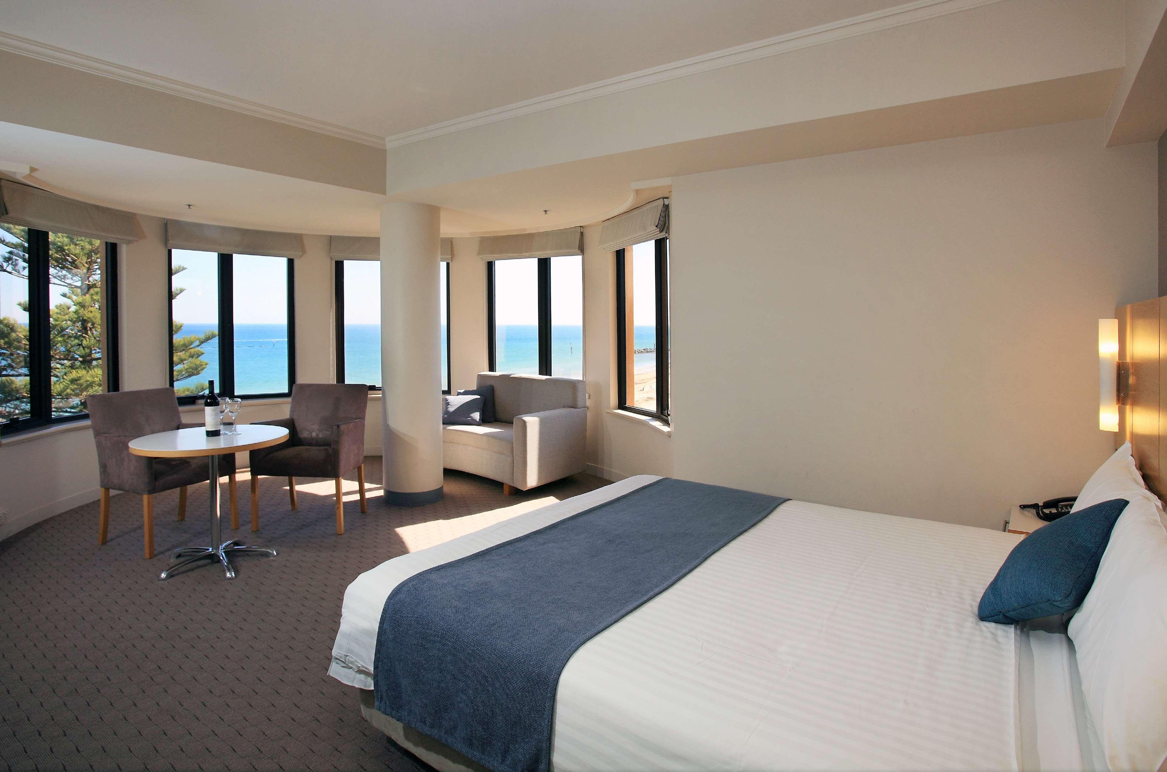 Отель Stamford Grand Adelaide Гленелг Экстерьер фото The photo shows a well-lit hotel room featuring large windows that offer a view of the ocean. The room is arranged with a comfortable bed covered in white linens and a navy blue throw blanket. There is a small table and chairs set up beside the windo