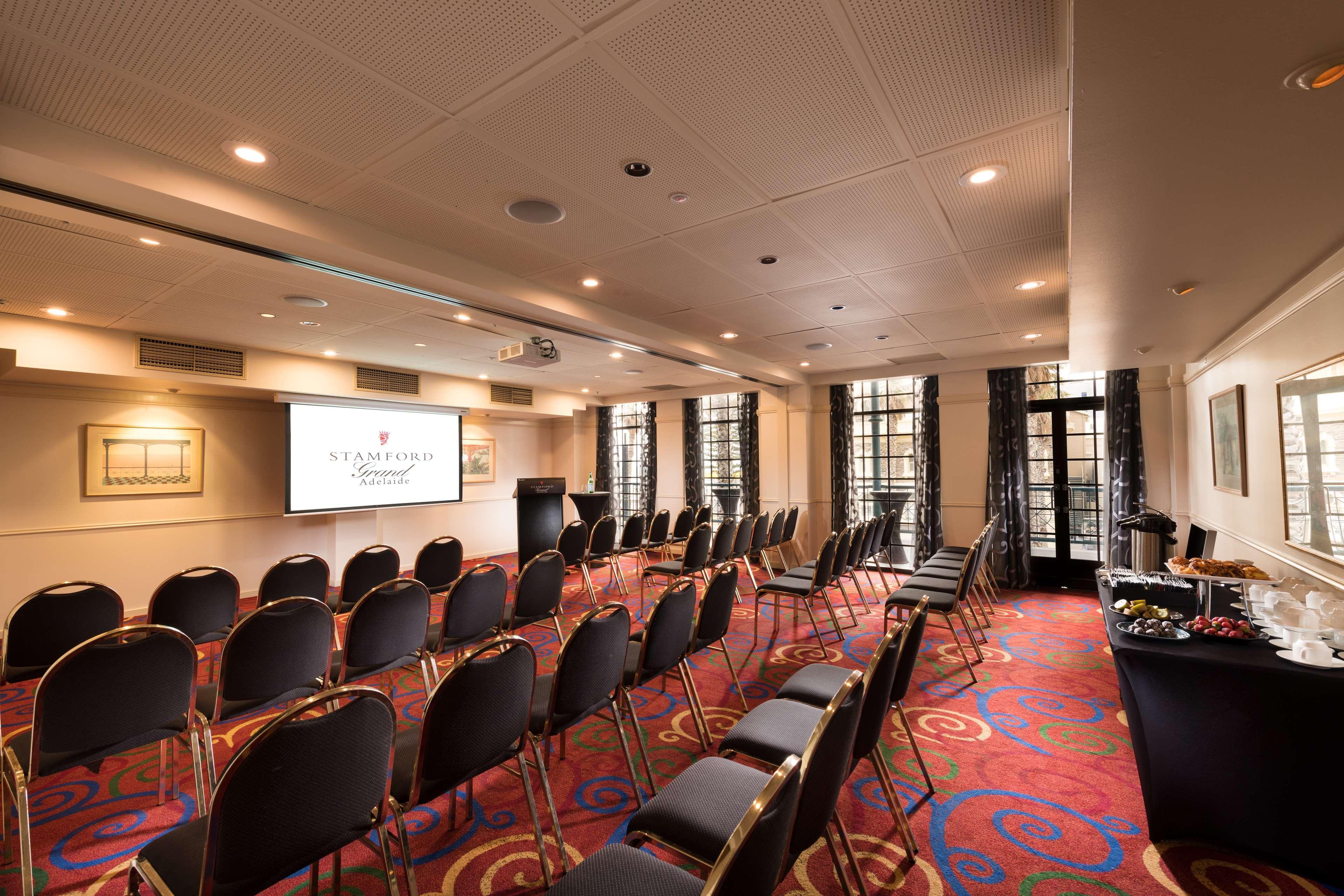 Отель Stamford Grand Adelaide Гленелг Экстерьер фото The photo shows a conference room set up for an event. There are rows of black chairs arranged in a semicircular formation facing a presentation screen at the front. The room features large windows that allow natural light to enter, and the flooring 
