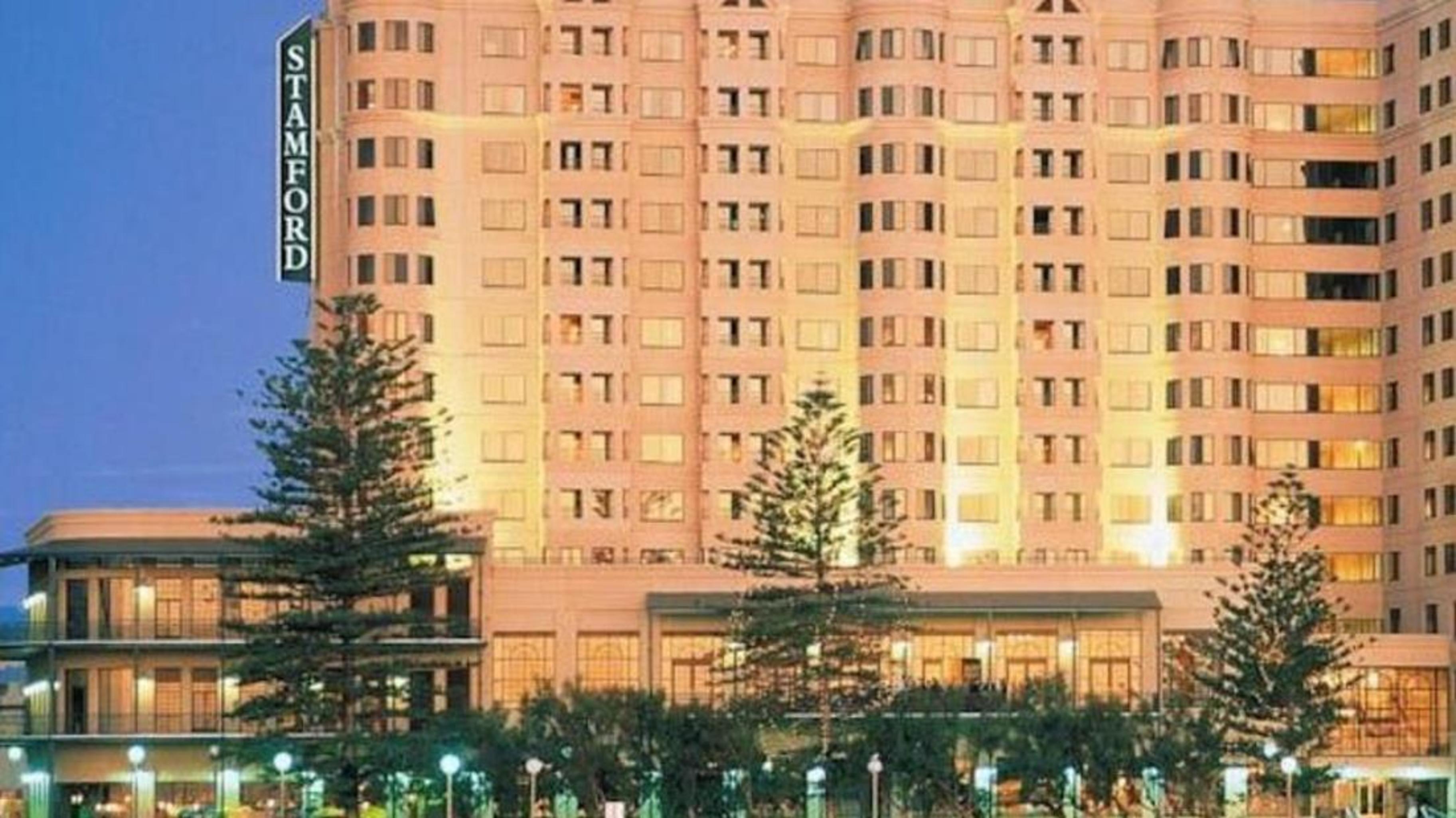 Отель Stamford Grand Adelaide Гленелг Экстерьер фото The photo shows a tall, modern building illuminated at night, likely a hotel, with the name "Stamford" prominently displayed on its side. Surrounding the building are several palm trees and landscaped areas, suggesting a tropical or coastal location.
