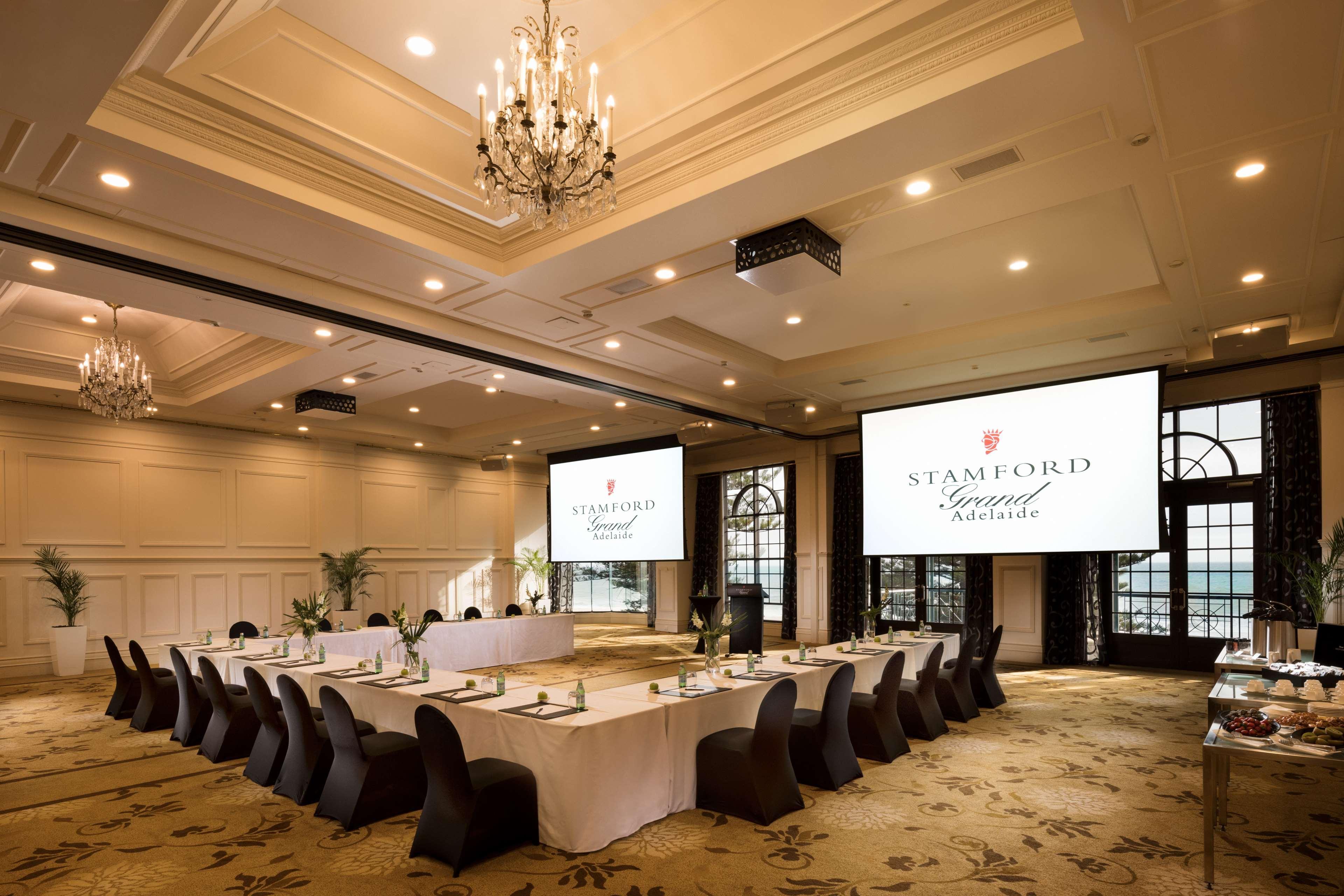 Отель Stamford Grand Adelaide Гленелг Экстерьер фото The photo shows a spacious and elegantly designed conference room. The room features a long table set for a meeting, surrounded by stylish chairs. Two large screens are positioned at the front, displaying the logo "Stamford Grand Melbourne." The ceil