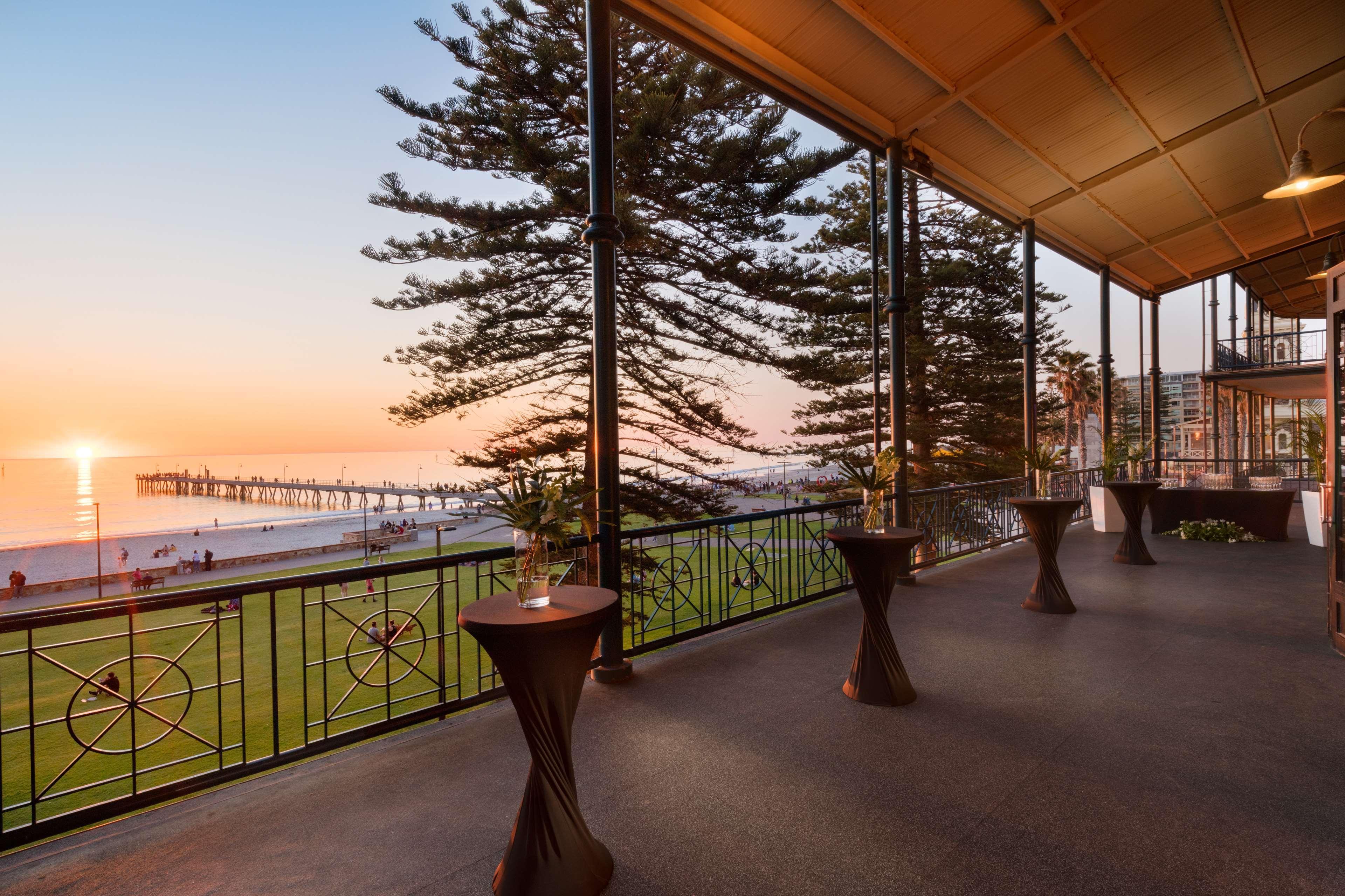 Отель Stamford Grand Adelaide Гленелг Экстерьер фото The photo shows a spacious indoor space with large windows overlooking a beach at sunset. The scene features a pier extending into the ocean and a horizon painted in warm colors from the setting sun. In the foreground, there are elegant tables with d