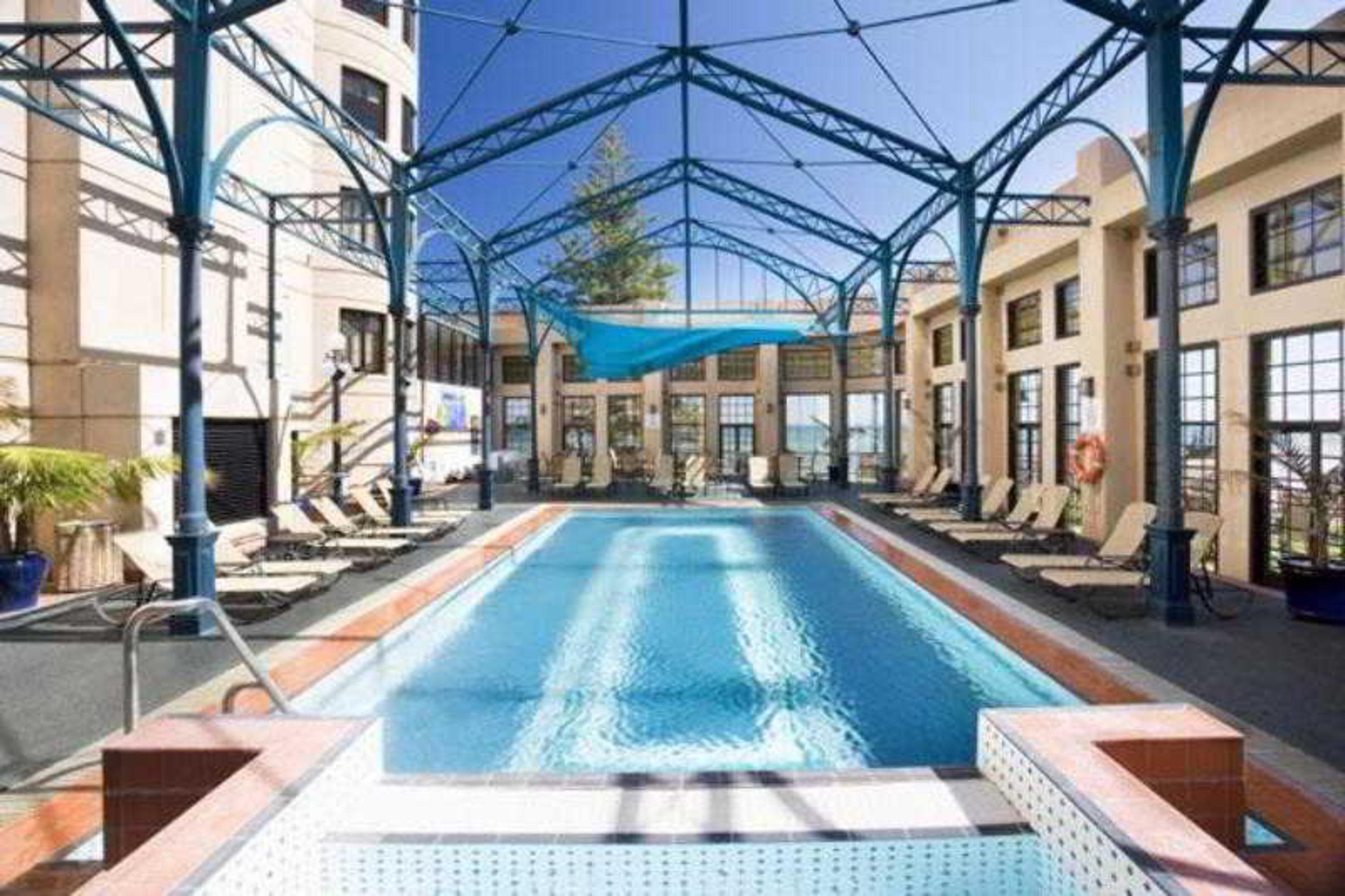 Отель Stamford Grand Adelaide Гленелг Удобства фото The photo shows an indoor swimming pool area. The pool is rectangular and features a blue cover above, supported by a metal frame. Surrounding the pool are several lounge chairs, and the space is well-lit with large windows allowing natural light to 