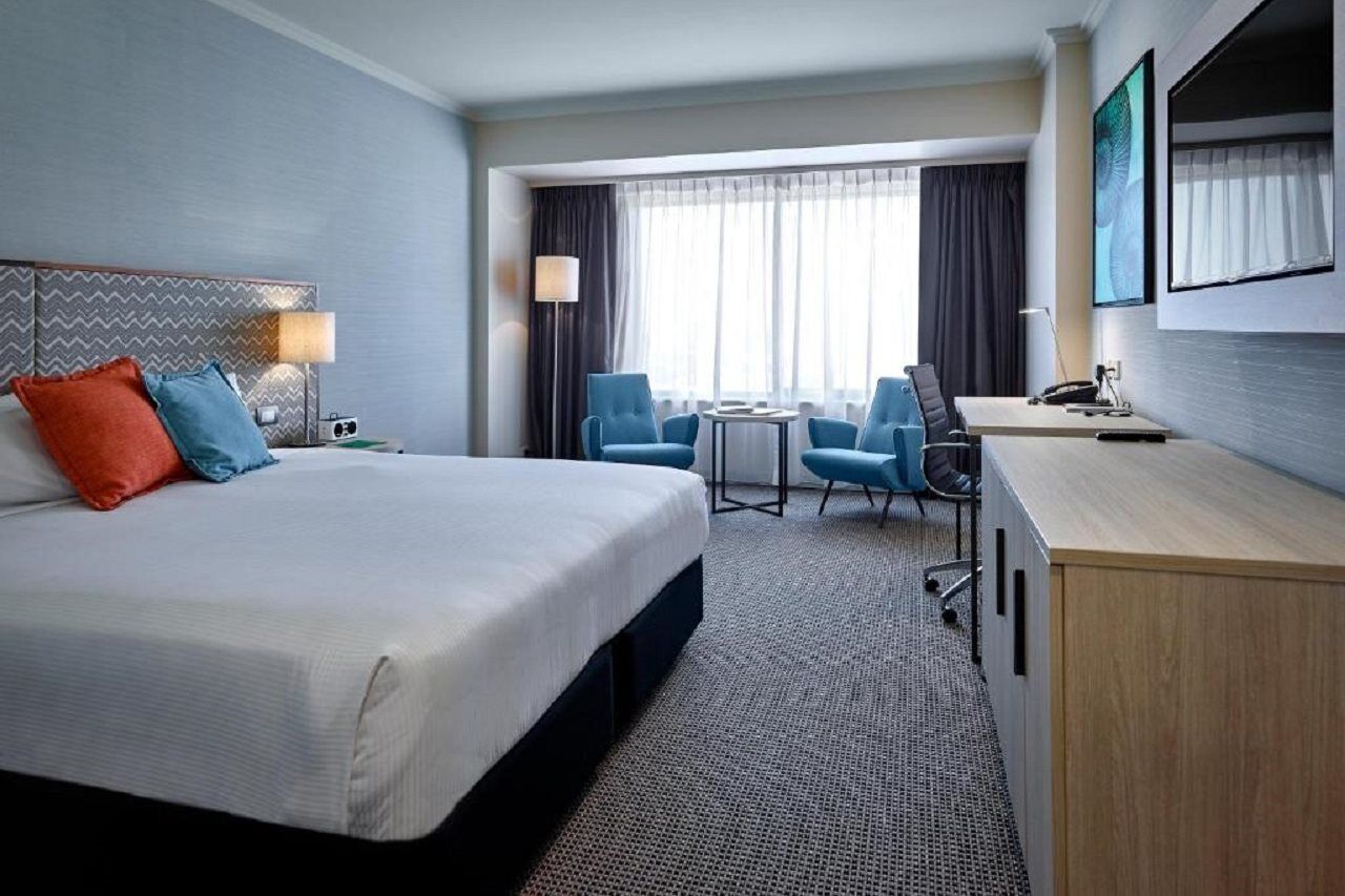 Отель Stamford Grand Adelaide Гленелг Экстерьер фото The photo shows a modern hotel room with a clean and minimalist design. In the center, there is a large, neatly made bed with white linens and two decorative pillows, one orange and one blue. To the right, there is a small wooden cabinet with a telev